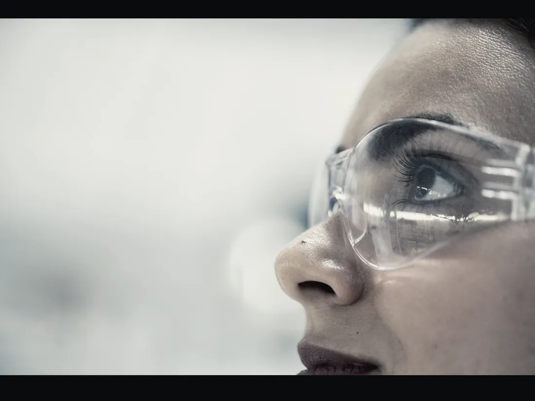 Technician in safety goggles closely inspecting work, seen in profile.